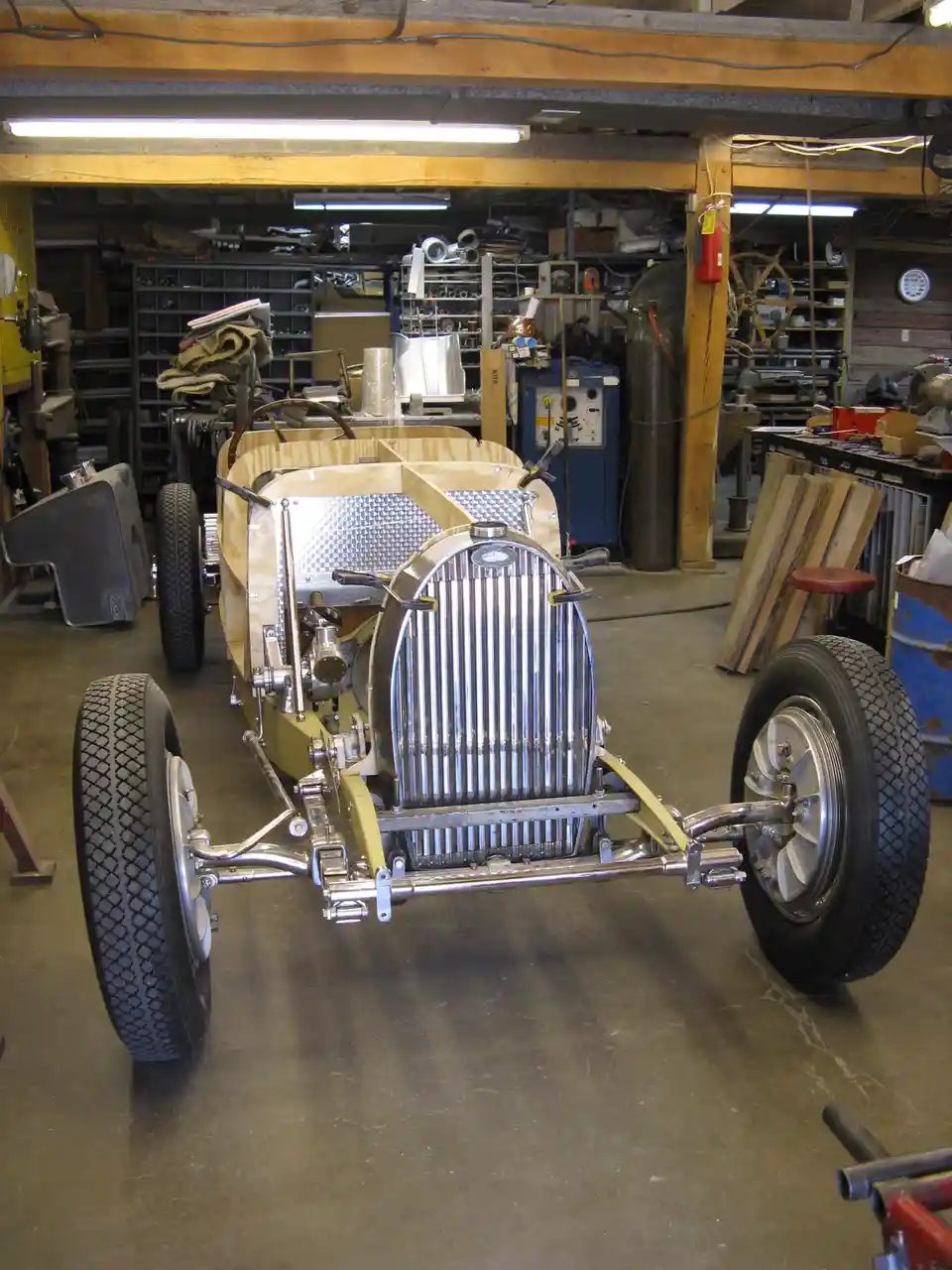 A partially assembled vintage-style race car sits in a cluttered workshop, featuring a shiny chrome grille, exposed chassis…