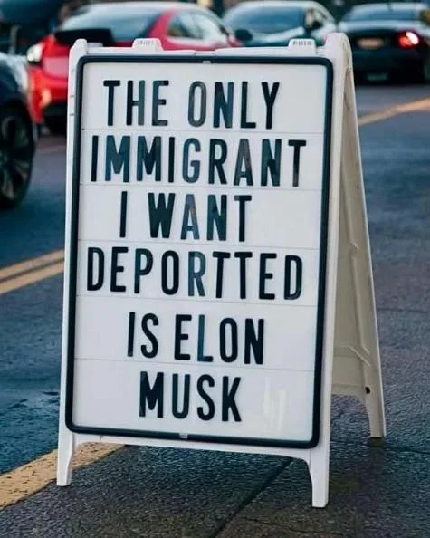 A white A-frame sign with black letter tiles displays the text 'THE ONLY IMMIGRANT I WANT DEPORTED IS ELON MUSK' in bold…