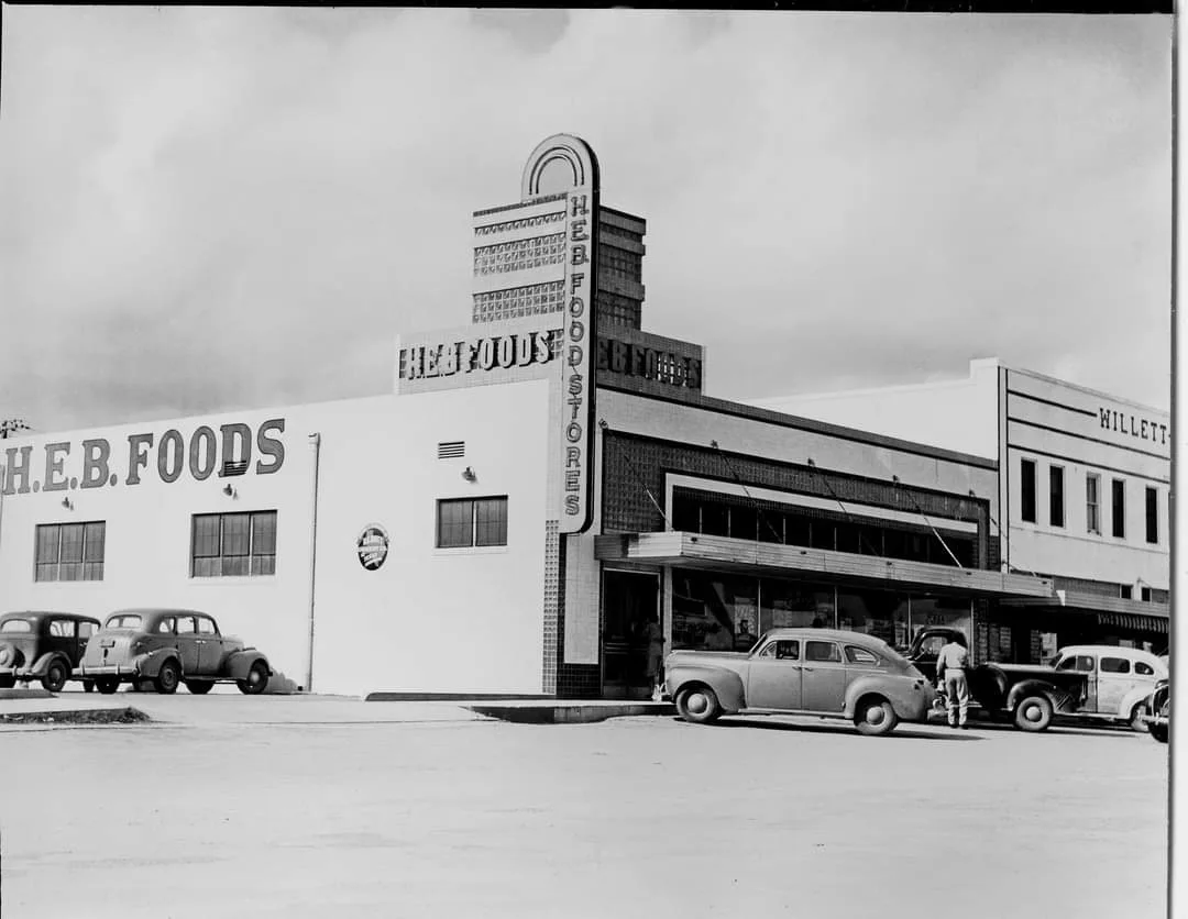 A black-and-white historical photo shows the exterior of an H.E.B. Foods store from the mid-20th century, with vintage cars…