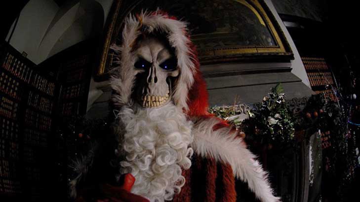 A skeletal figure dressed in a Santa Claus costume with a white beard and red hat stands in a dimly lit, ornate room…