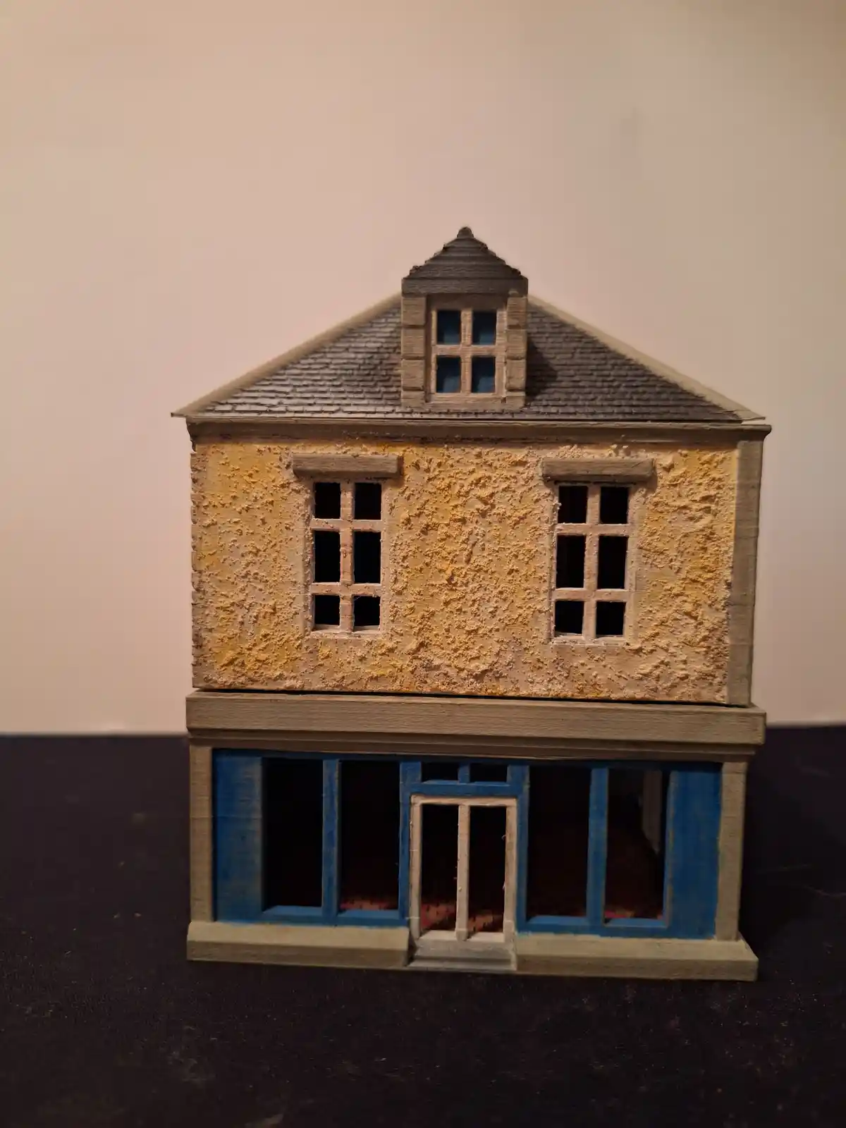 A 3d-printed miniature house model with a yellow textured upper floor, blue-painted lower floor, and a grey roof with a…
