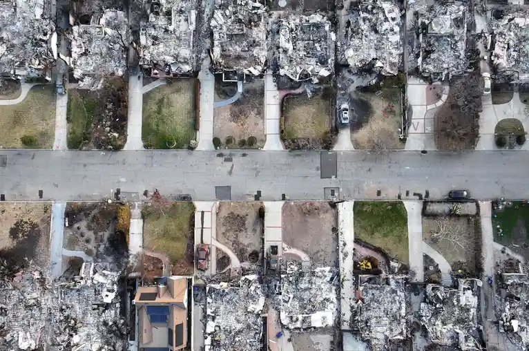 An aerial view shows a neighborhood devastated by fire, with most houses reduced to charred rubble and debris. One house…
