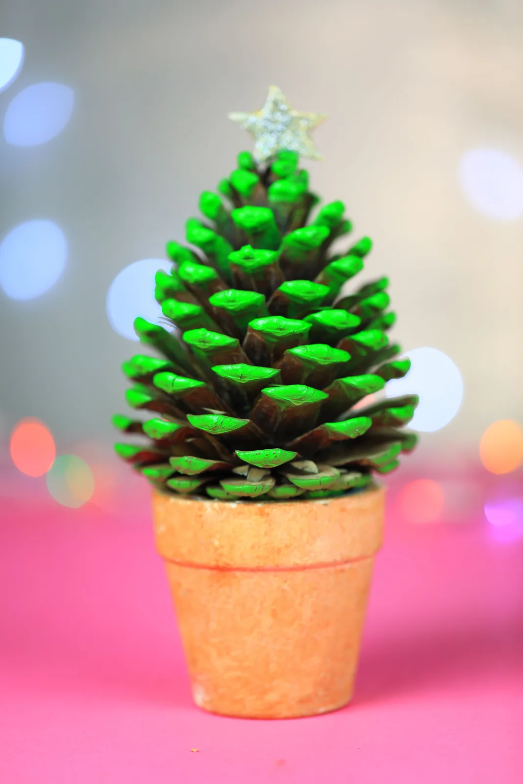 A pine cone with its scales painted bright green and topped with a glittery star sits in a small terracotta pot on a pink…