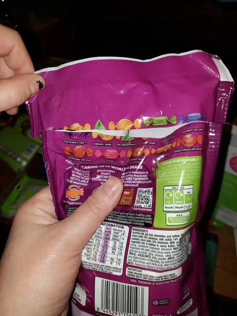 A person holds a purple Quality Street candy bag, pointing at the nutritional information section with their thumb. The back…
