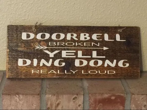A weathered wooden sign mounted on a brick wall and beige stucco reads 'DOORBELL BROKEN' with an arrow pointing right…