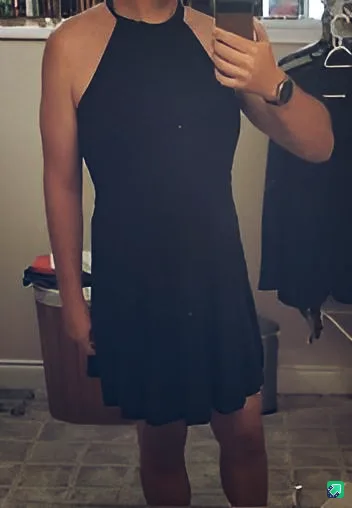 A person stands in front of a mirror holding a phone to take a selfie, wearing a simple black halter-neck dress. The…