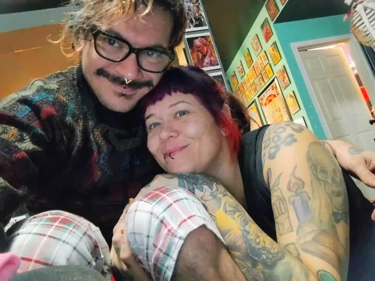 A selfie of a man with glasses, a mustache, and a nose ring, and a woman with purple hair and extensive tattoos, both…