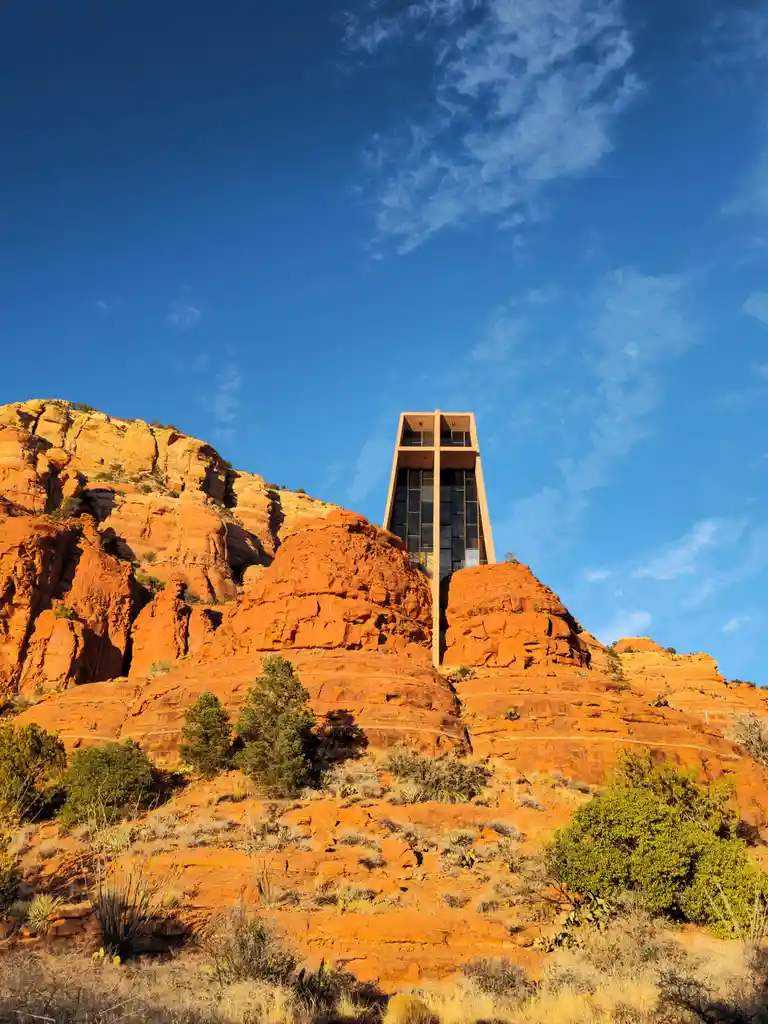 a modern, rectangular church building with glass windows is perched atop a steep, red rock cliff in a desert landscape under…
