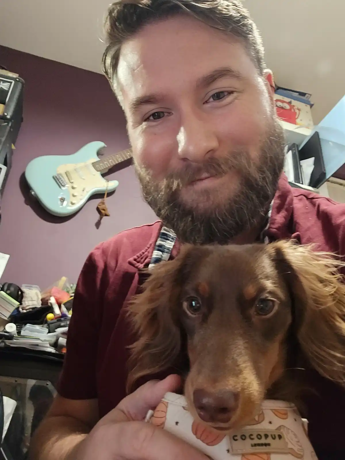 A man with a beard and a red shirt holds a long-haired dachshund wearing a 'COCOPUP LONDON' bib. The dog looks directly at…