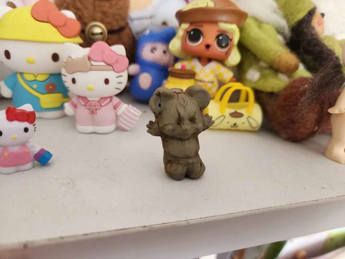 A small, gray, textured bear figurine stands alone on a shelf, surrounded by colorful, well-cared-for toys like Hello Kitty…