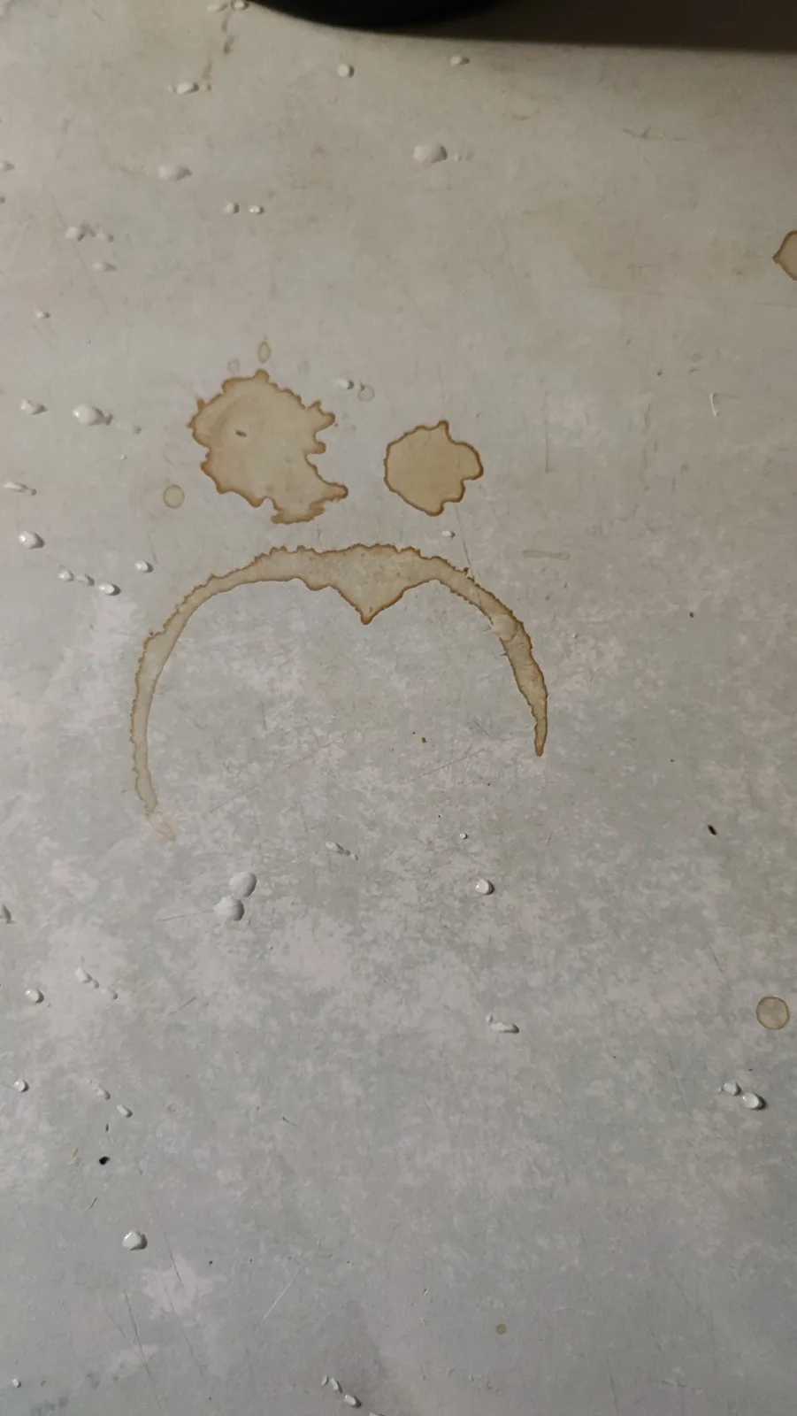 A close-up of a light-colored surface, likely a countertop or table, shows two brown liquid stains forming eyes and a…