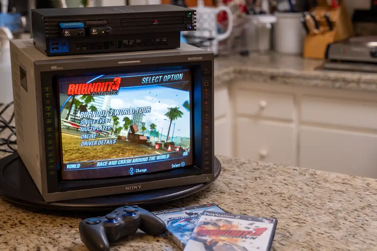 A PlayStation 2 console sits atop a small Sony CRT television displaying the 'Burnout 3: Takedown' main menu, with game…