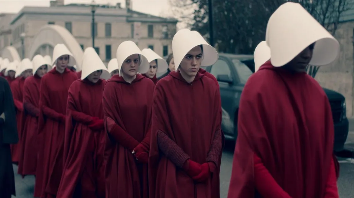 A line of women in red cloaks and white bonnets from the TV show 'The Handmaid's Tale' stand in formation outdoors, looking…