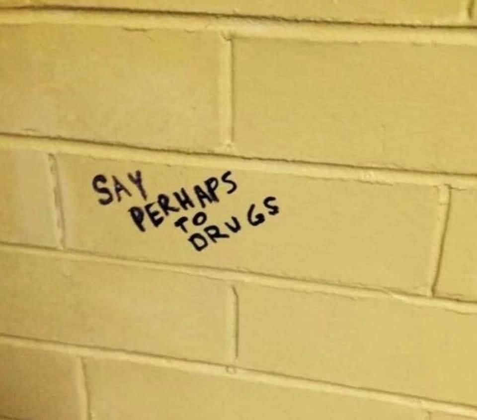 A close-up photo of a pale yellow brick wall with handwritten text in black marker on one brick that reads 'SAY PERHAPS TO…