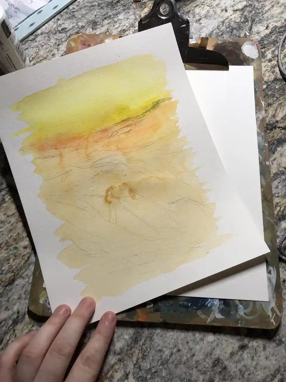 A hand holds a watercolor painting on paper, showing a rough, abstract portrait with yellow and orange washes and faint…