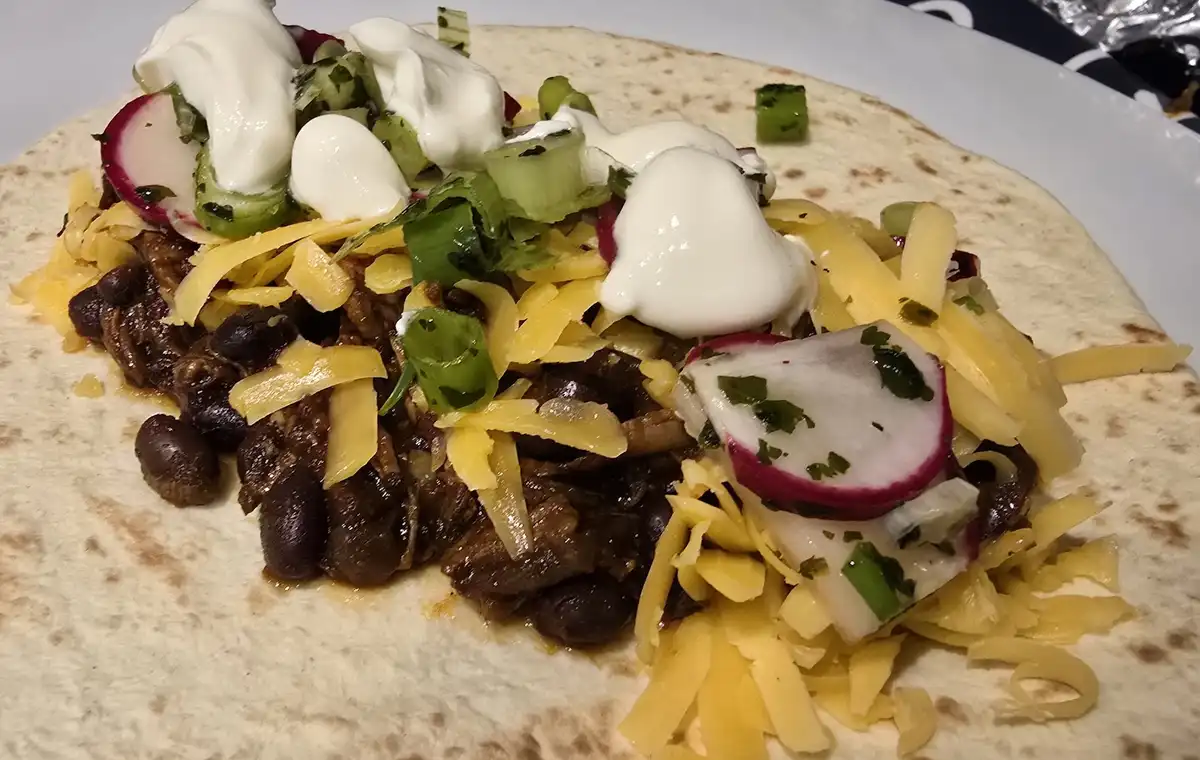 a close-up photo of a large, open-faced taco on a white plate, filled with shredded yellow cheese, black beans, meat, sliced…