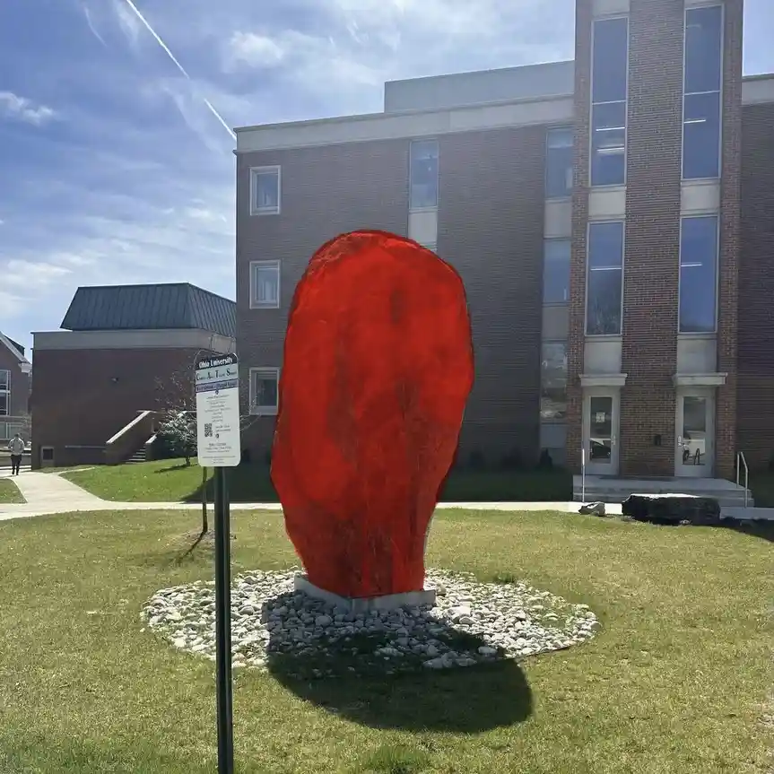 A large, bright red, roughly oval-shaped sculpture resembling a giant rock stands on a grassy campus lawn surrounded by…