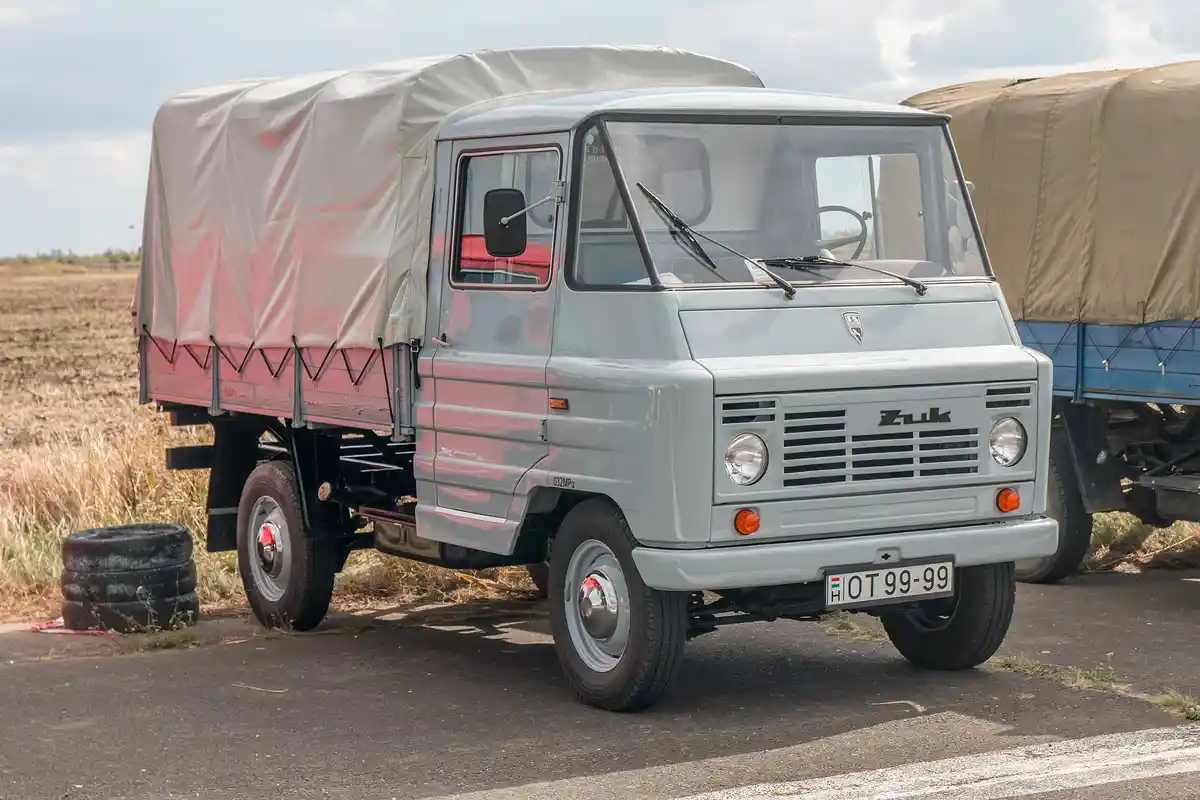 A light gray vintage ZUK truck with a canvas-covered cargo bed is parked outdoors, its license plate reads 'HOT 99-99'. The…