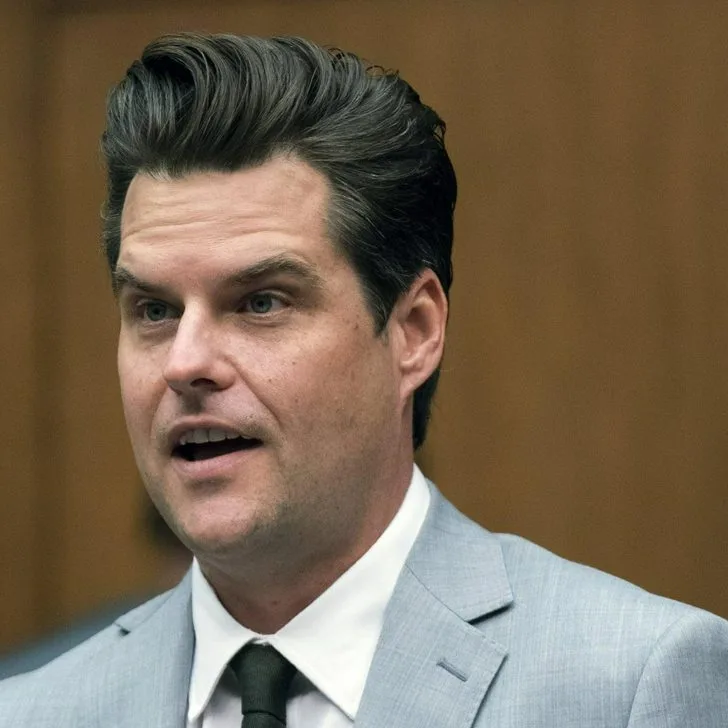 a man with dark hair and blue eyes, wearing a light gray suit, white shirt, and dark tie, is captured mid-speech with his…