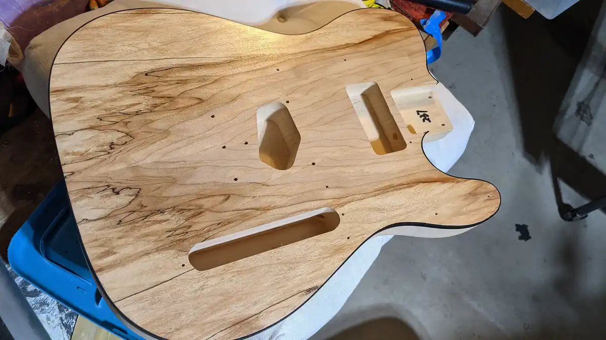 A close-up photo shows the unfinished wooden body of an electric guitar, resting on a white cloth. The natural wood grain…