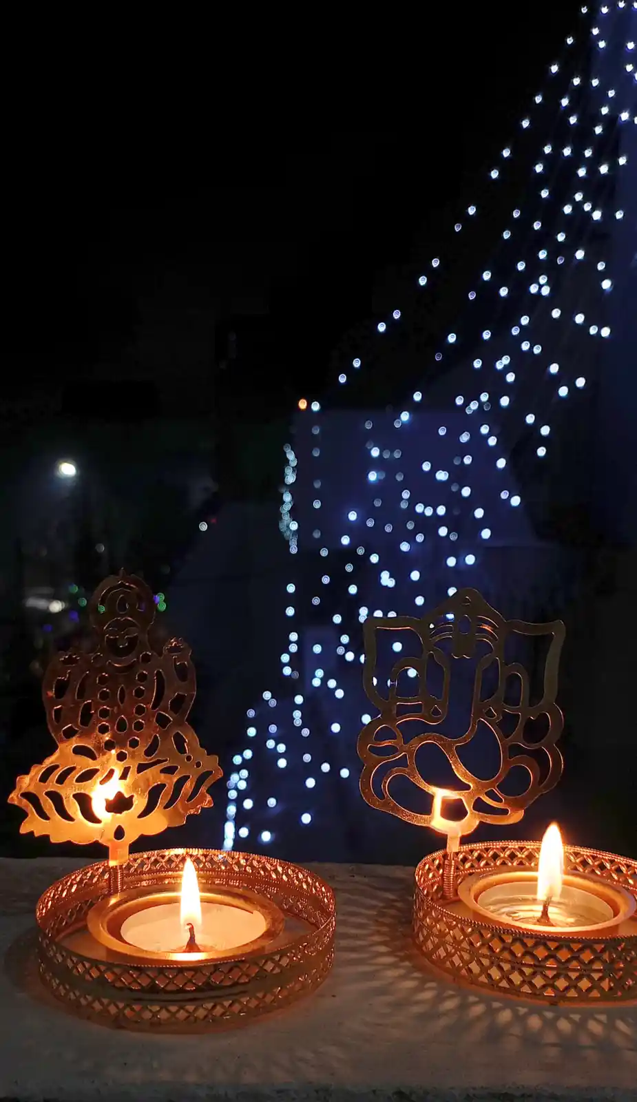 Two ornate, lit tea light candles in decorative holders shaped like Hindu deities Ganesha and Krishna are in the foreground…