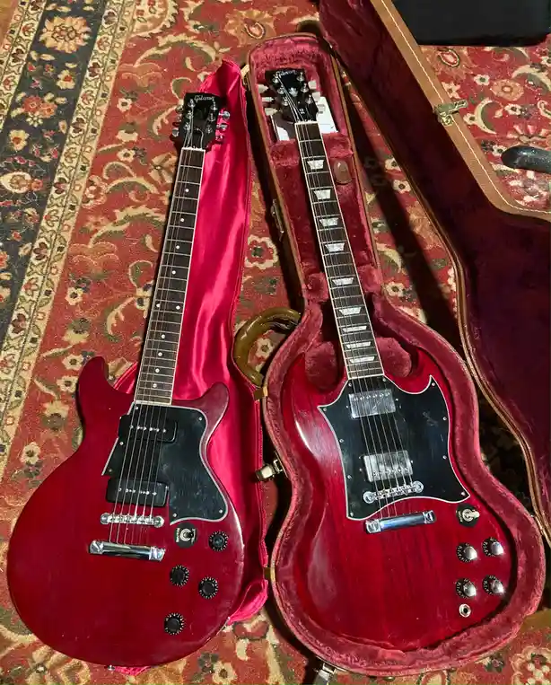 Two identical red Gibson electric guitars are displayed side-by-side on a patterned rug. One guitar rests outside its open…