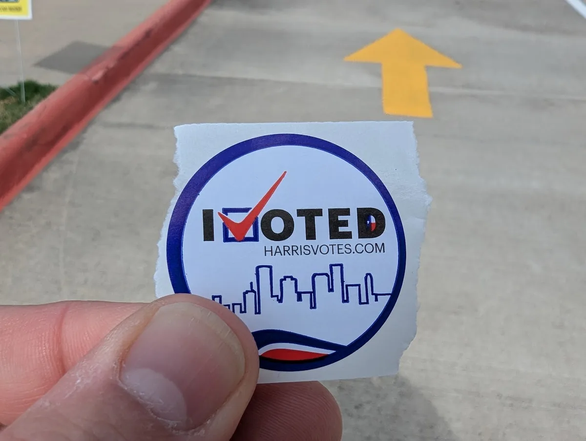 A hand holds a torn 'I VOTED' sticker from Harris County, Texas, featuring a red checkmark over the 'I', a city skyline…