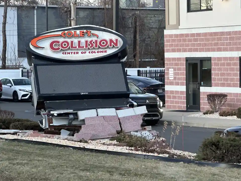 A large, damaged sign for 'Cole's Collision Center of Colonie' is visibly toppled over, with its base broken and bricks…