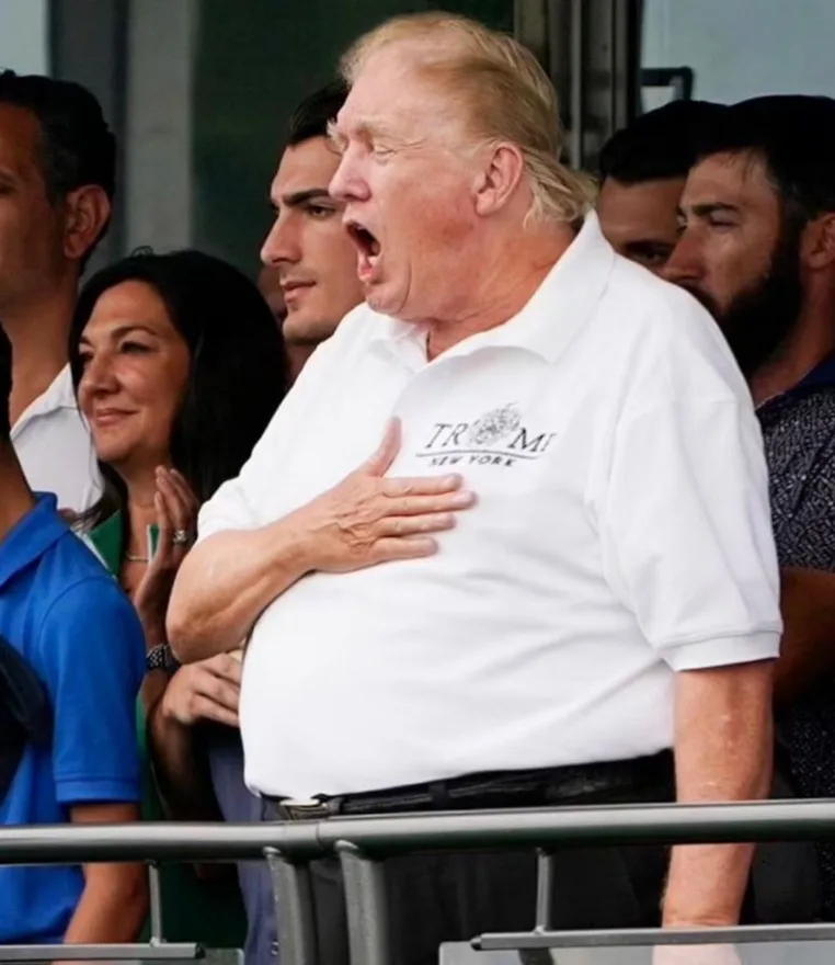A man in a white polo shirt with 'TRUMP NEW YORK' embroidered on the chest stands with his hand over his heart and mouth…
