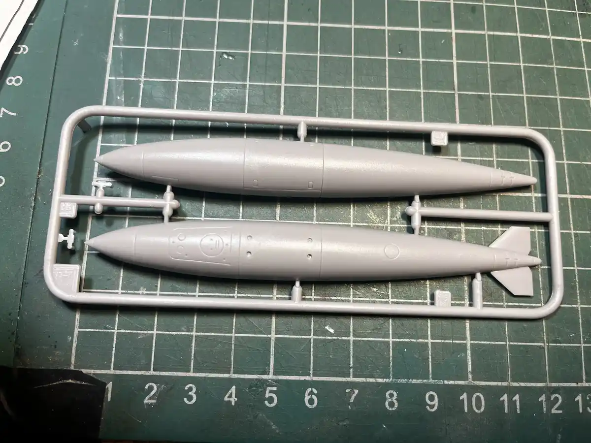 A close-up photo shows two unpainted grey plastic model missile parts still attached to their sprue, resting on a green…