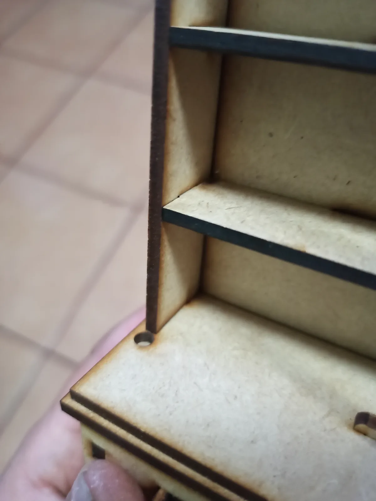 A close-up photo shows a hand holding a small, unfinished wooden shelf unit with laser-cut edges. The shelf has multiple…