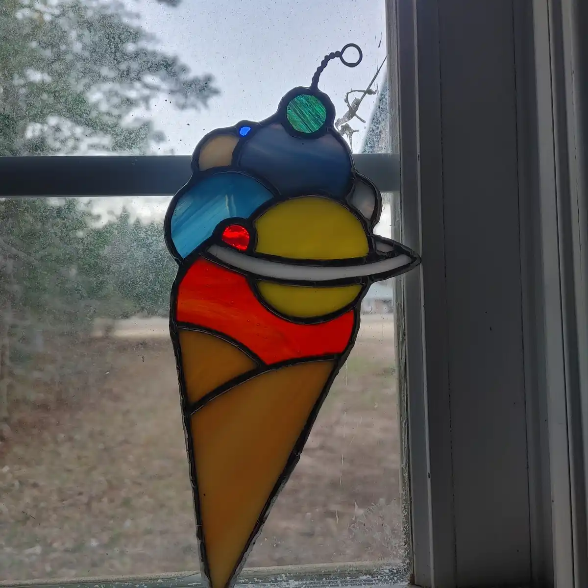 A stained glass window hanging depicts a colorful ice cream cone with multiple scoops, featuring red, yellow, blue, and…