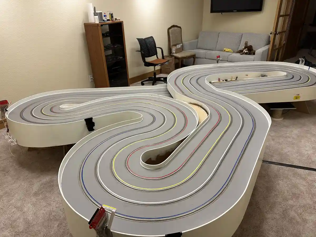 A large, custom-built slot car racing track with multiple colorful lanes is installed in a living room, taking up most of…