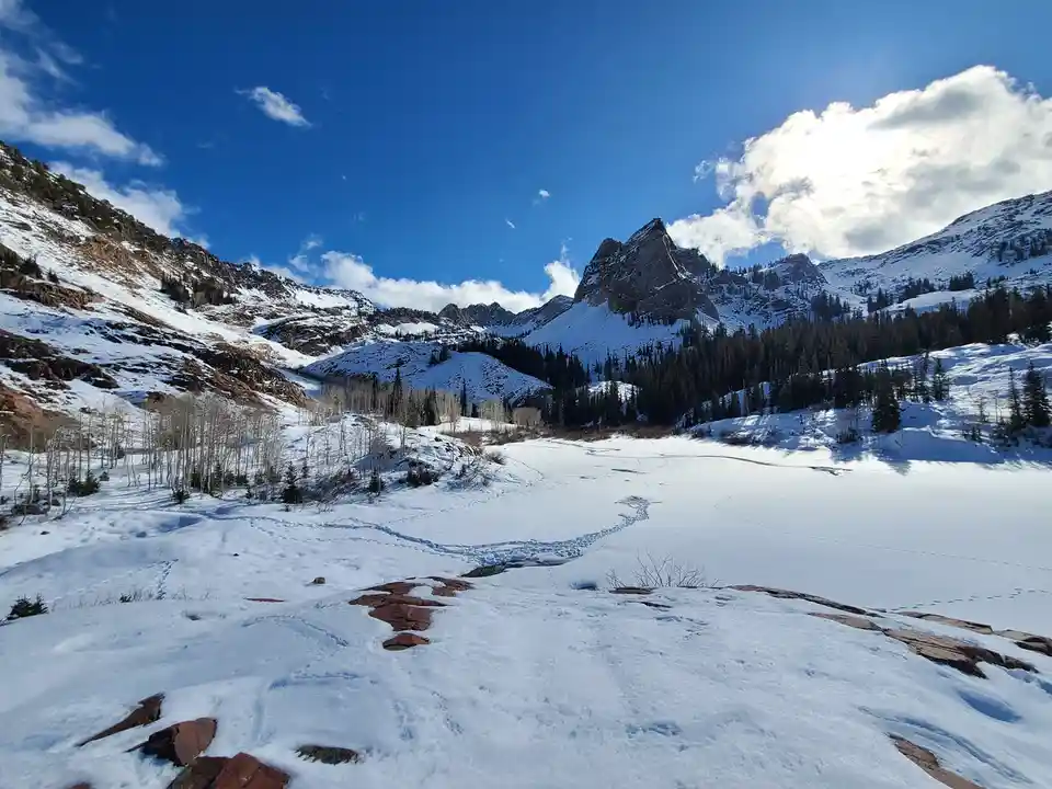 A scenic winter landscape shows snow-covered mountains under a bright blue sky with scattered clouds. A frozen lake…