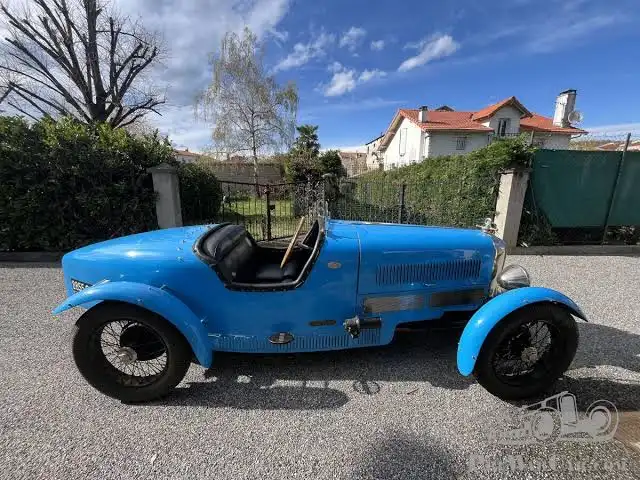 a bright blue vintage open-top sports car with a wooden steering wheel and black leather seat is parked on a gravel…