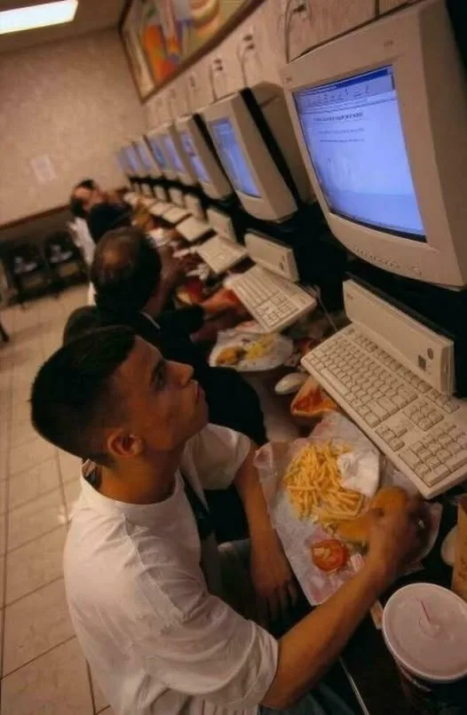 A young man sits at a row of old beige computer terminals in a fast-food restaurant, eating fries and a burger while looking…