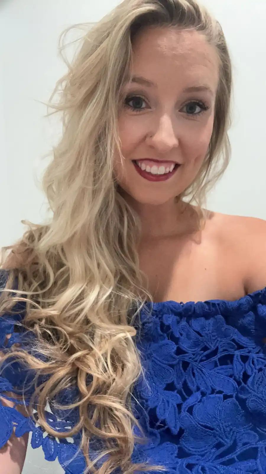 A smiling woman with long blonde hair wearing a blue lace off-the-shoulder top poses for a selfie against a plain white…