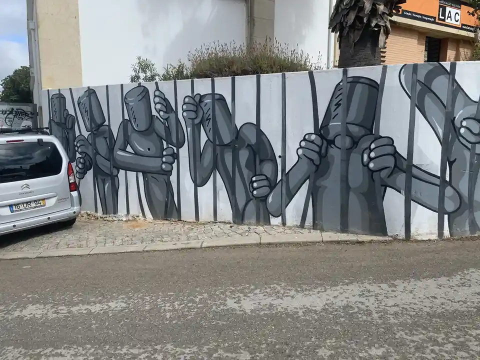 A street mural depicts several gray, headless cartoon figures behind black bars, gripping the bars with their hands. A…