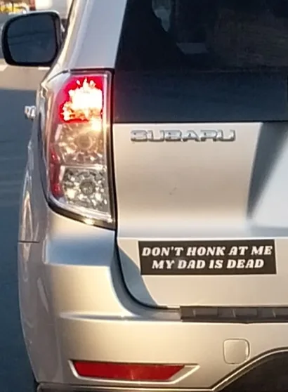 The rear of a silver Subaru vehicle shows a black bumper sticker with white text reading 'DON'T HONK AT ME MY DAD IS DEAD'…