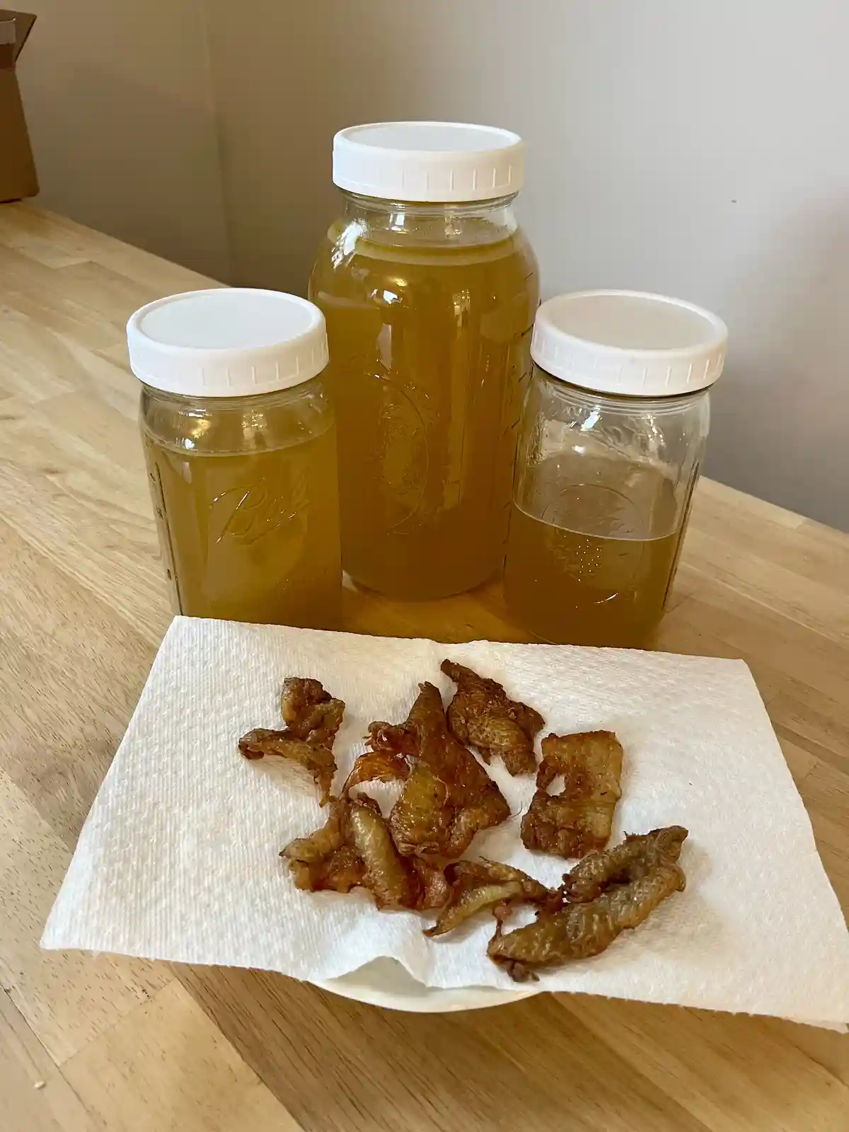 Three glass jars filled with golden-brown grease sit on a wooden table, with a white paper towel on a plate in front holding…