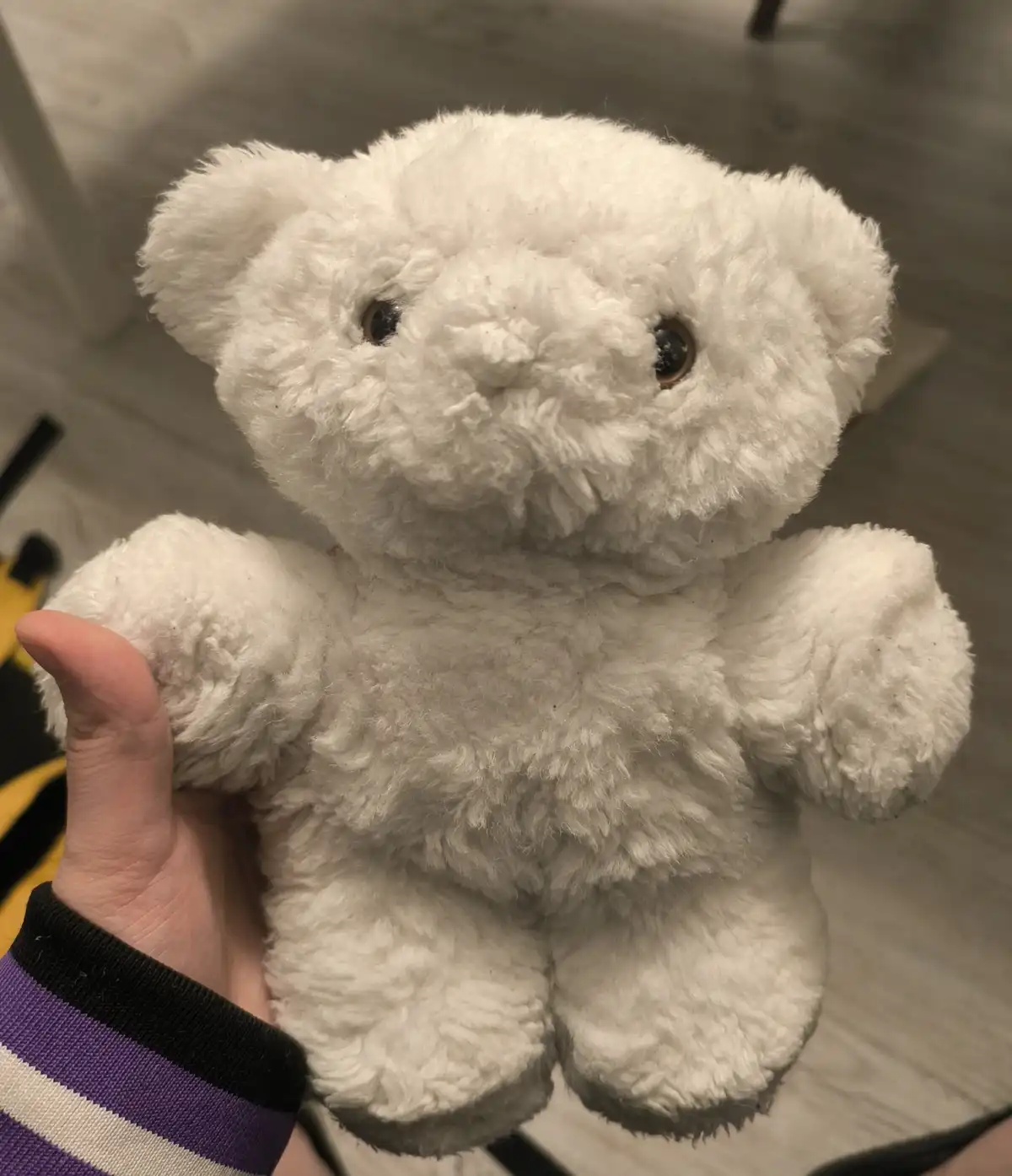 A person holds a white, fluffy stuffed teddy bear with dark button eyes, presenting it to the camera. The bear's fur appears…