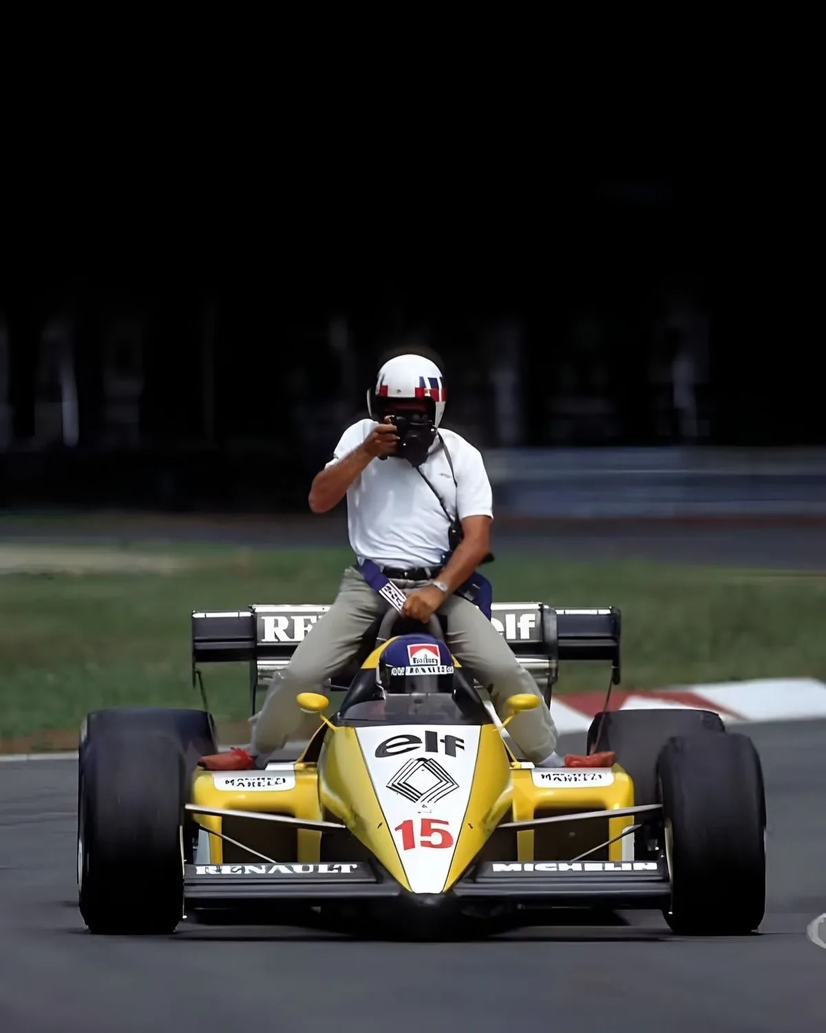 A man in a white helmet and shirt is sitting on the rear wing of a yellow and black Formula 1 race car, holding a camera up…
