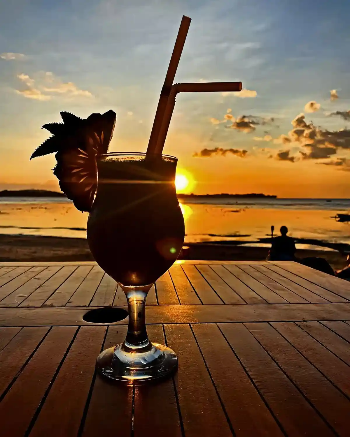 A tropical cocktail in a stemmed glass with a straw and pineapple garnish sits on a wooden table, silhouetted against a…