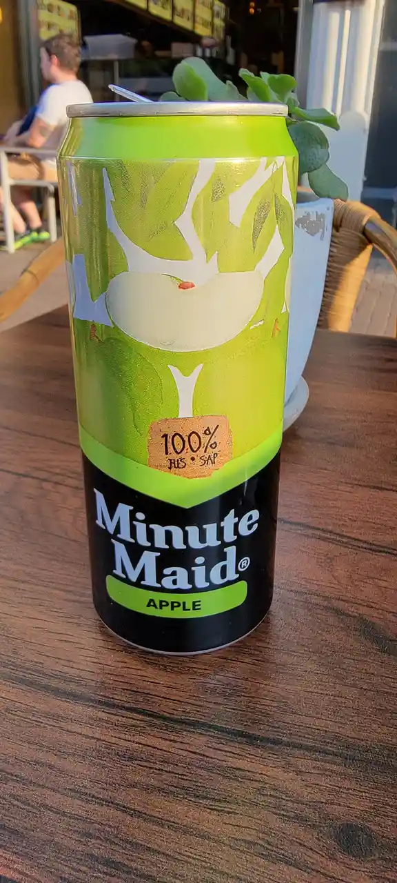 A close-up photo of a green and black Minute Maid Apple juice can sitting on a wooden table, with the text '100% JUS * SAP'…