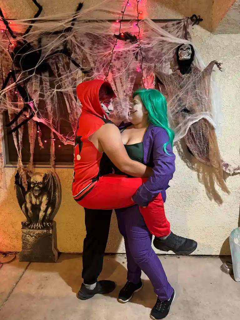 Two people dressed as Harley Quinn and the Joker embrace in front of a Halloween-decorated wall with spiderwebs, red lights…
