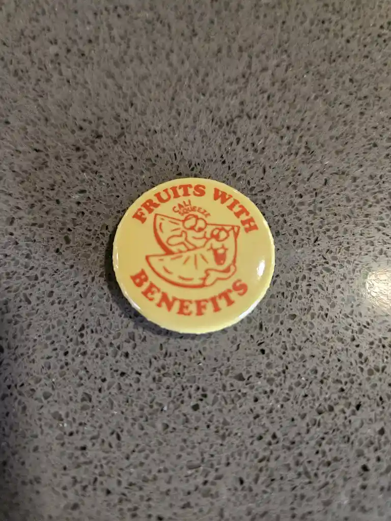 A yellow circular button with red text and a cartoon citrus slice logo sits on a speckled gray countertop. The text reads…