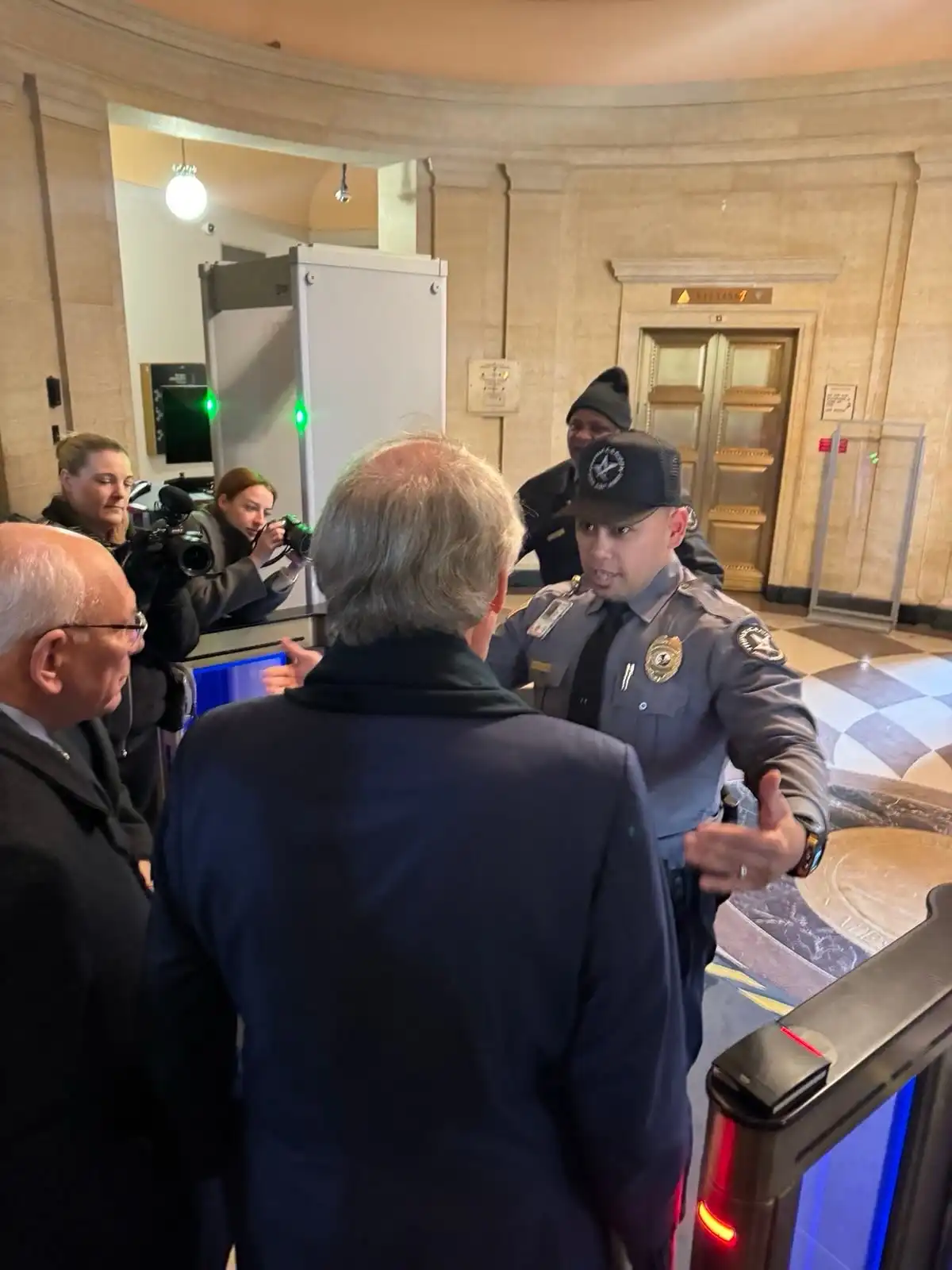 A security guard in a grey uniform and cap is gesturing and speaking to a man in a dark suit from behind, while another man…