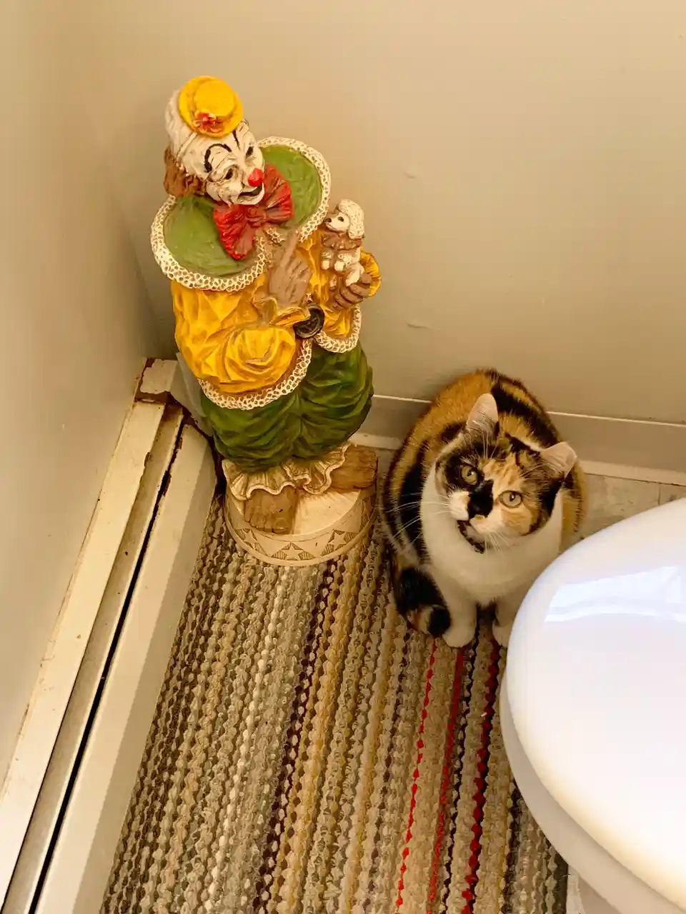 A calico cat sits on a patterned bathroom rug next to a colorful clown statue, looking up with wide eyes. The clown statue…