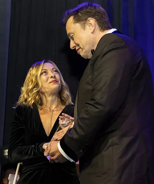 A man in a black suit leans down to speak to a woman in a black velvet dress holding a glass award; she looks up at him with…