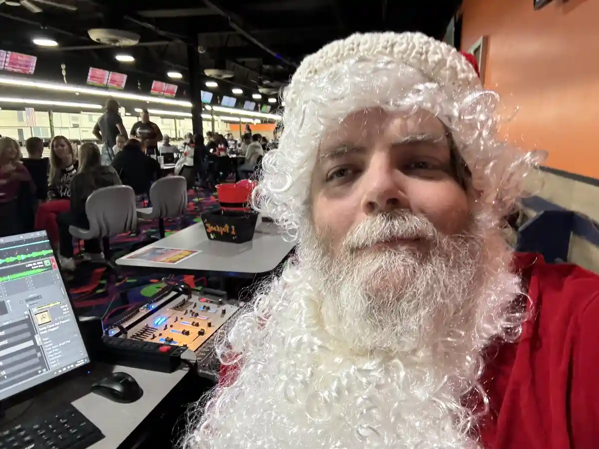 A man dressed as Santa Claus with a white wig and beard sits in front of a sound mixing console and laptop in what appears…
